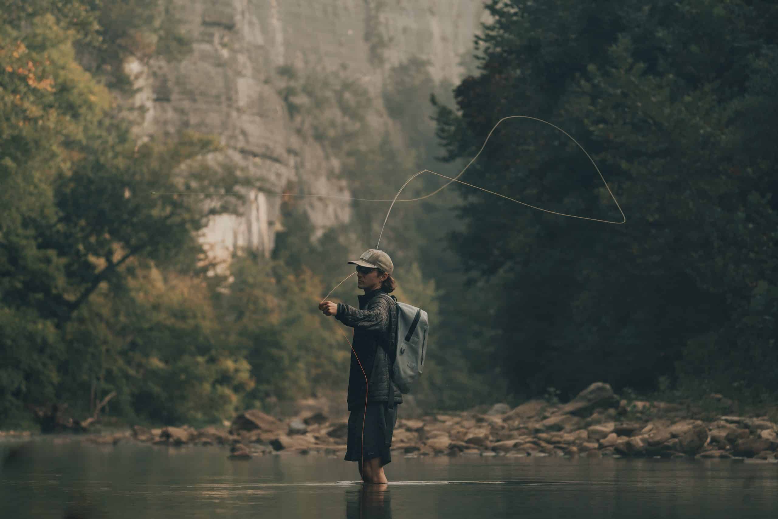 Man with backpack casting fly fishing pole below sunny trees Stock